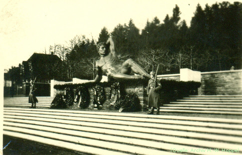 Foto: Eine liegende, übergroße, nackte männliche Figur mit einem Lorbeerkranz auf einem Sockel. Vor dem Sockel Ehrenkränze. Rechts und links daneben Soldaten als Ehrenwache.
