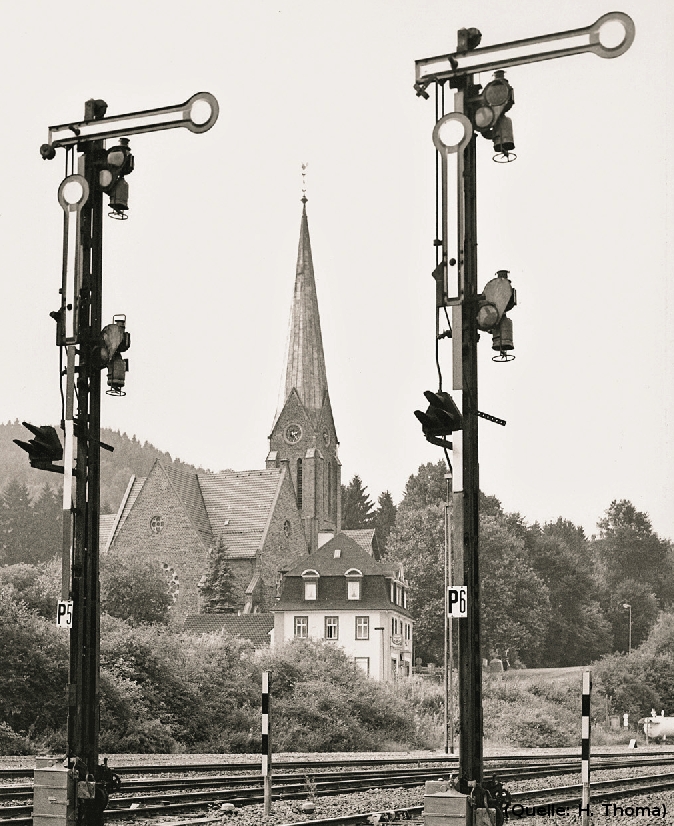 Foto: Zwei Bahnsignale stehen auf 'HALT'. dazwischen ist in der Ferne eine Kirche zu sehen.