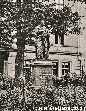 Foto: Ein Denkmal mit der Figur Wilhelms des I. auf einem Sockel. Dahinter ein Haus.