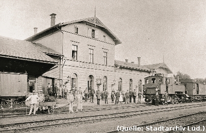 Foto: Ein Bahnhof. Davor Personen und ein Zug mit einer Dampflok.