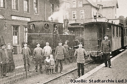 Foto: Ein Bahnhof. Davor Personen und ein Zug mit einer Dampflok.