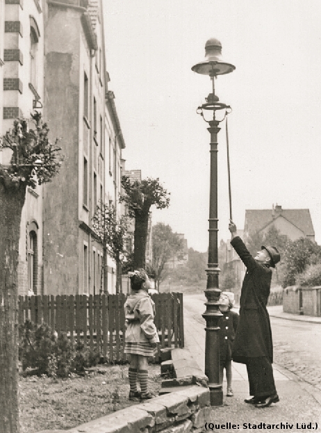 Foto: Ein Mann mit einem stockähnlichem Gegenstand bedient eine ca. 4 Meter hohe Gaslaterne an einer Strasse. Zwei Kinder sehen zu.