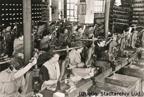 Foto: Blick in eine Fabrikhalle. Frauen sitzen an langen Tischen und bedienen Handpressen.