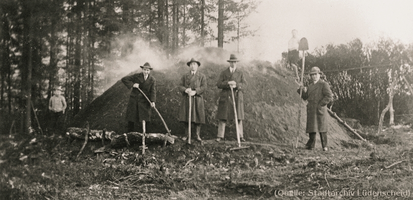 Foto: Von 1930: Eine Gruppe Männer mit Schaufeln stehen vor einem rauchenden Holzkohlenmeiler.
